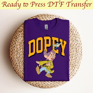 May include: Purple t-shirt with a yellow graphic of Dopey from Disney's Snow White and the Seven Dwarfs. Dopey is walking and holding a bird in his hand. The text "Dopey" is in a yellow block letter font.