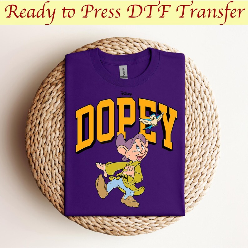 May include: Purple t-shirt with a yellow graphic of Dopey from Disney's Snow White and the Seven Dwarfs. Dopey is walking and holding a bird in his hand. The text "Dopey" is in a yellow block letter font.