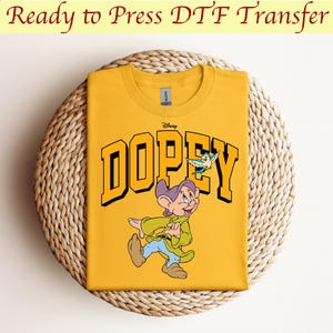 May include: Yellow t-shirt with a graphic of Dopey from Disney's Snow White and the Seven Dwarfs. The graphic features Dopey walking with a bird flying above him. The text "Disney" and "DOPEY" are printed in black.