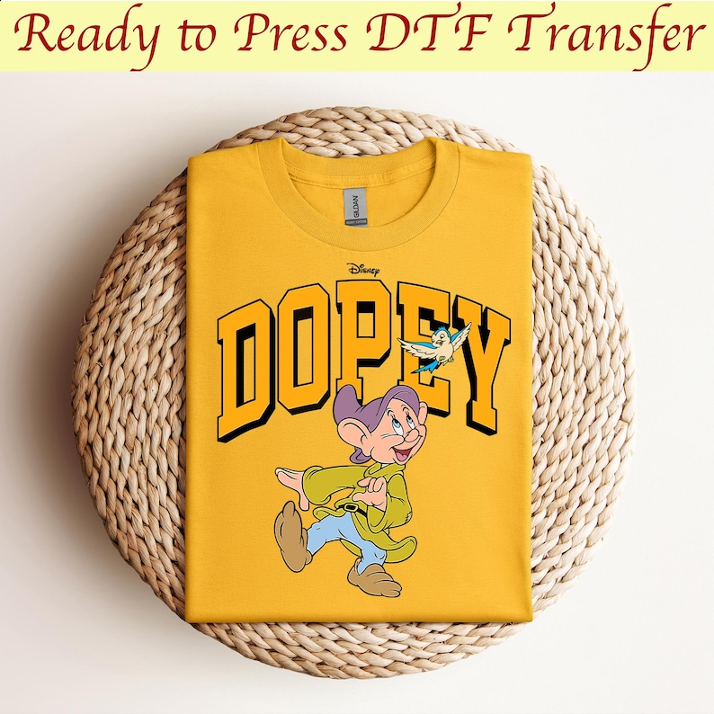 May include: Yellow t-shirt with a graphic of Dopey from Disney's Snow White and the Seven Dwarfs. The graphic features Dopey walking with a bird flying above him. The text "Disney" and "DOPEY" are printed in black.