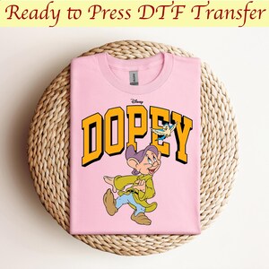 May include: Pink t-shirt with a yellow graphic of Dopey from Disney's Snow White and the Seven Dwarfs. The graphic includes the word "Dopey" in yellow letters and a small blue bird.