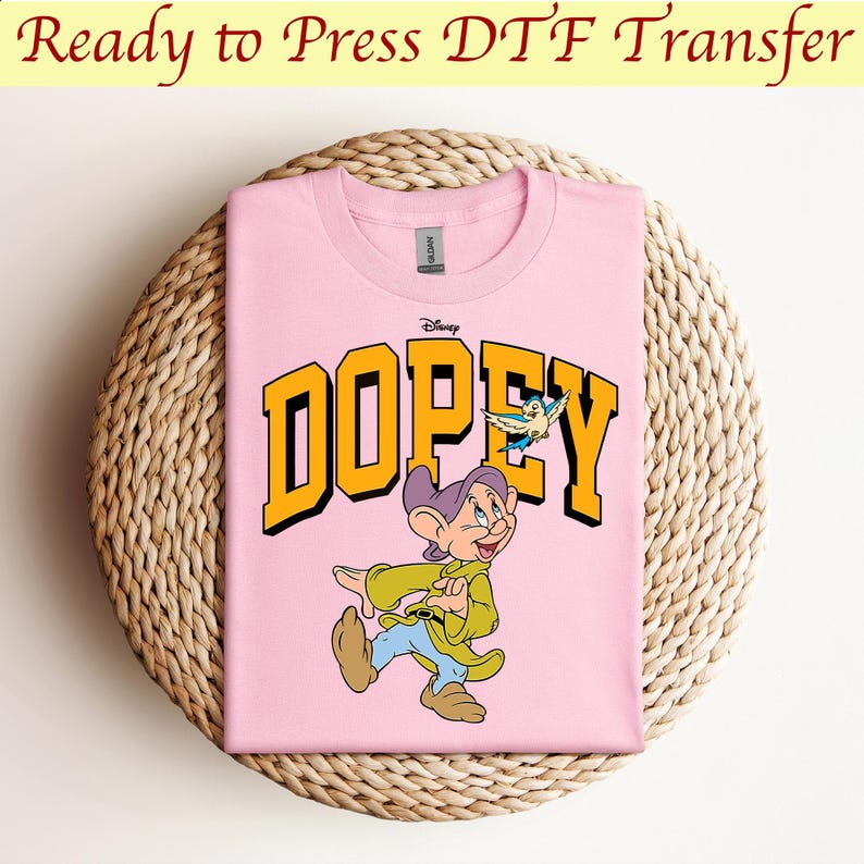May include: Pink t-shirt with a yellow graphic of Dopey from Disney's Snow White and the Seven Dwarfs. The graphic includes the word "Dopey" in yellow letters and a small blue bird.