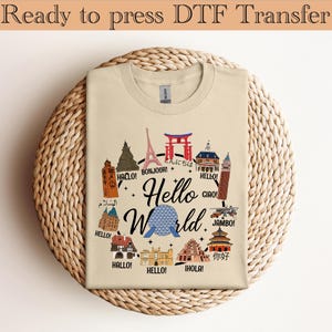 Op de afbeelding: Een beige t-shirt met de tekst "Hello World" in een gestileerd lettertype, omringd door illustraties van bezienswaardigheden en begroetingen in verschillende talen. Het t-shirt ligt op een ronde, geweven ondergrond.