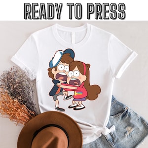 Op de afbeelding: Wit T-shirt met een grafische print van twee cartoonfiguren, een jongen en een meisje, met de tekst "READY TO PRESS" bovenaan.