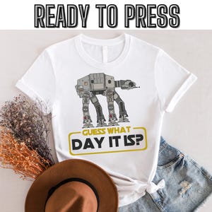 Puede incluir: Camiseta blanca con un gráfico de un caminante AT-AT de Star Wars y el texto "Adivina qué día es?" impreso en negro y dorado.