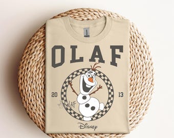 Puede incluir: Una camiseta de color beige con un gráfico en blanco y negro de Olaf de Frozen. El gráfico incluye el texto "Olaf" en letras negras, un círculo a cuadros alrededor de Olaf y el texto "Disney" debajo del círculo.