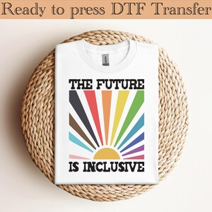 Puede incluir: Camiseta blanca con el texto "THE FUTURE IS INCLUSIVE" en negro. El diseño presenta un sol radiante colorido con rayos de colores arcoíris. La camiseta se muestra sobre una superficie circular tejida.