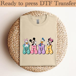 Könnte beinhalten: Ein beiges T-Shirt mit Rundhalsausschnitt und einem Grafikdruck des Wortes MAMA, bei dem jeder Buchstabe durch eine Disney-Figur dargestellt wird: Daisy Duck, Mickey Mouse, Donald Duck und Minnie Mouse, die jeweils auf einem bunten gepunkteten Buchstaben sitzen.