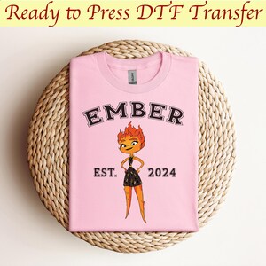 May include: Pink t-shirt with a black and white graphic of a cartoon character with flames for hair. The text "EMBER EST. 2024" is printed on the shirt.