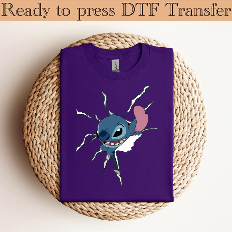 K&ouml;nnte beinhalten: Ein lila T-Shirt mit einem Cartoon-Charakter, der durch den Stoff zu brechen scheint. Der Charakter ist blau mit einem rosa Ohr und wei&szlig;en Z&auml;hnen. Der Text "Ready to press DTF Transfer" steht oben.