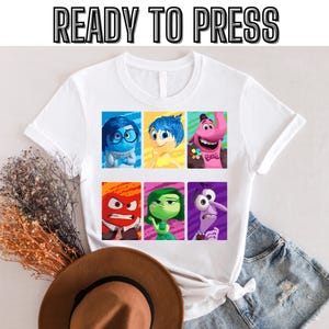 以下が含まれることがあります： 映画「インサイド・ヘッド」のキャラクターがプリントされた白いTシャツ。キャラクターは6つの正方形のグリッドに配置され、各キャラクターは異なる色の正方形に入っています。キャラクターは、喜び、悲しみ、恐怖、怒り、嫌悪、そしてビング・ボンです。画像の上部に「READY TO PRESS」というテキストが印刷されています。