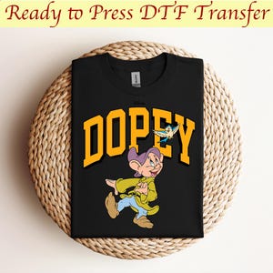 May include: Black t-shirt with a yellow "DOPEY" graphic featuring a cartoon image of Dopey from Snow White and the Seven Dwarfs. Dopey is wearing a purple hat and a yellow jacket and is walking with a blue bird flying above him.