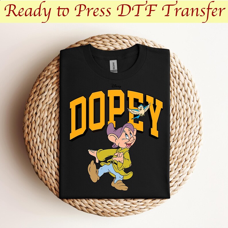 May include: Black t-shirt with a yellow "DOPEY" graphic featuring a cartoon image of Dopey from Snow White and the Seven Dwarfs. Dopey is wearing a purple hat and a yellow jacket and is walking with a blue bird flying above him.