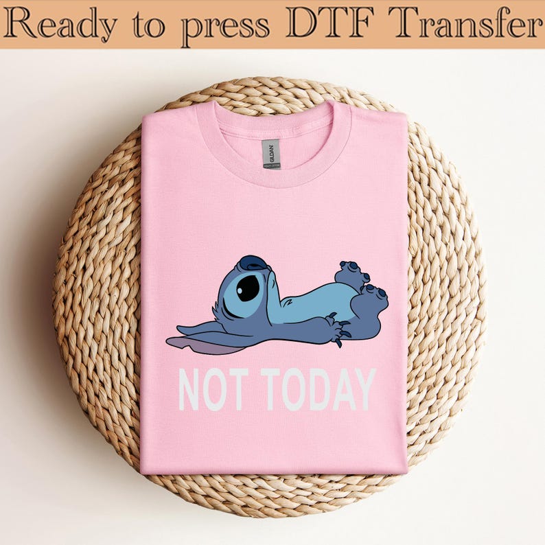 K&ouml;nnte beinhalten: Ein hellrosa T-Shirt mit einer Grafik einer blauen au&szlig;erirdischen Figur, die auf dem R&uuml;cken liegt, darunter der Text NOT TODAY in wei&szlig;en Gro&szlig;buchstaben. Das T-Shirt hat einen Rundhalsausschnitt und ein sichtbares Markenetikett am Kragen.