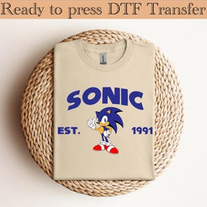 May include: A tan t-shirt featuring a blue graphic of Sonic the Hedgehog with the word "SONIC" above the character. Below the character are the words "EST. 1991". The shirt is folded and placed on a woven, circular surface.