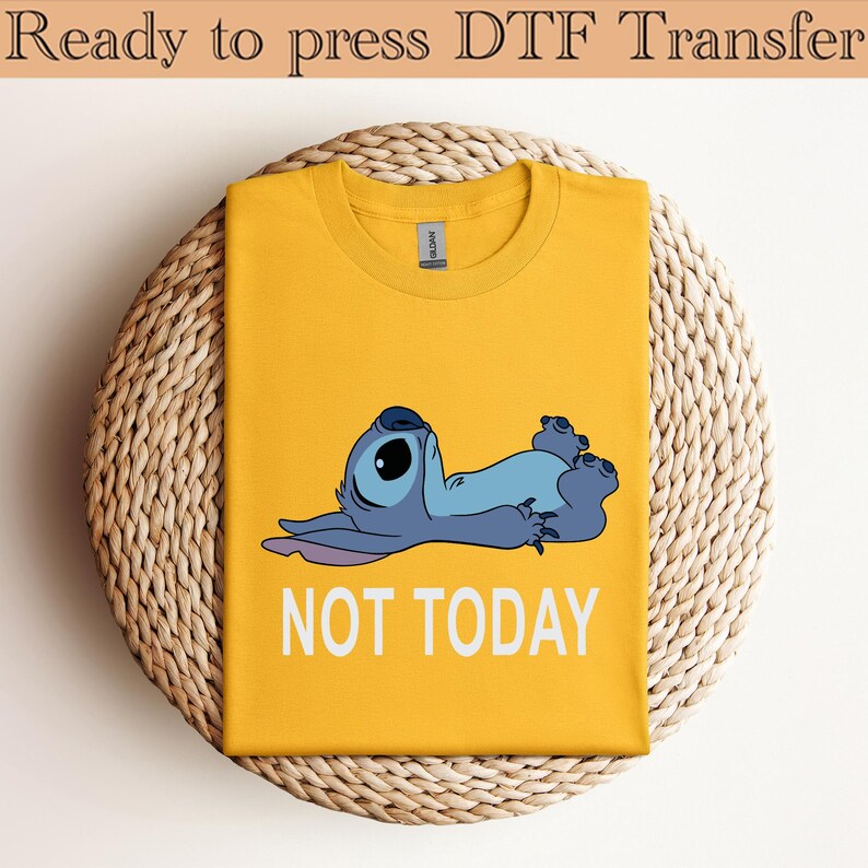 K&ouml;nnte beinhalten: Ein senfgelbes T-Shirt mit Rundhalsausschnitt und einer Grafik eines blauen Alien-Charakters, der auf dem R&uuml;cken liegt, mit dem darunter gedruckten wei&szlig;en Text NOT TODAY. Das T-Shirt hat ein klassisches Design mit kurzen &Auml;rmeln.