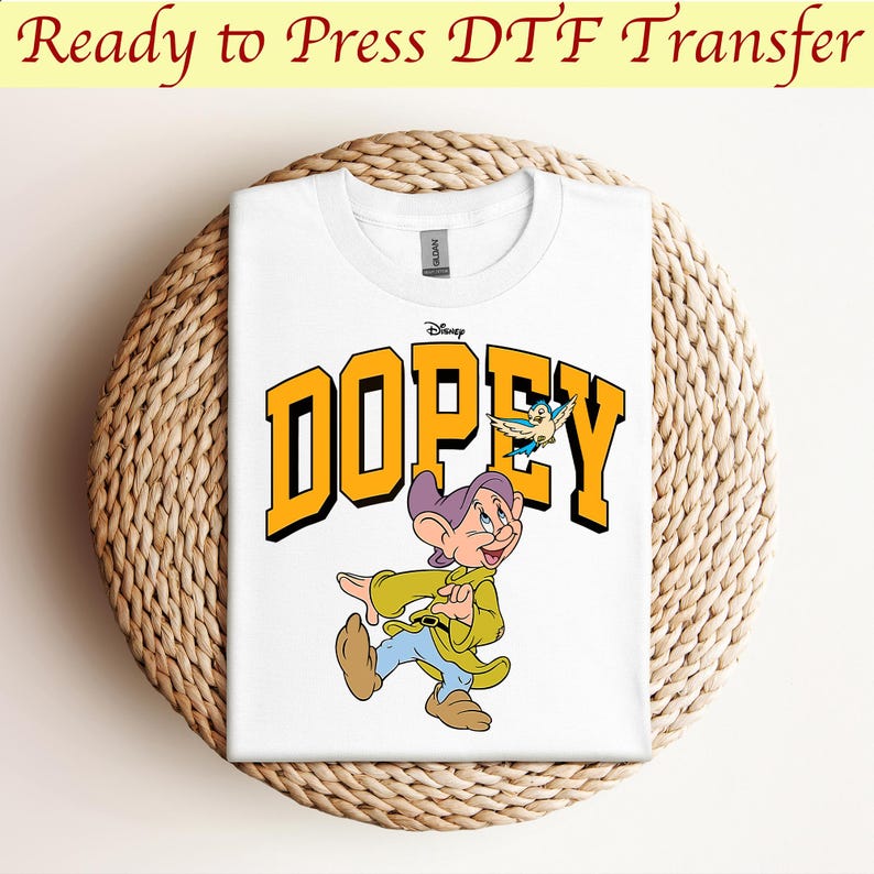 May include: White t-shirt with a yellow and blue graphic of Dopey from Disney's Snow White and the Seven Dwarfs. The graphic includes the text "Dopey" in yellow letters and a small blue bird.