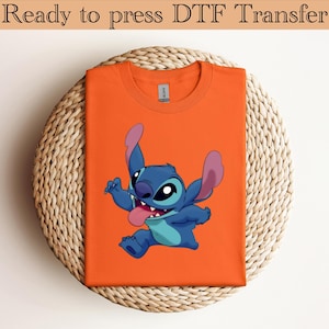 May include: Orange t-shirt featuring a cartoon character, Stitch, with blue fur, large ears, and a pink tongue. The text "Ready to press DTF Transfer" is at the top. The shirt is folded and placed on a woven, circular surface.