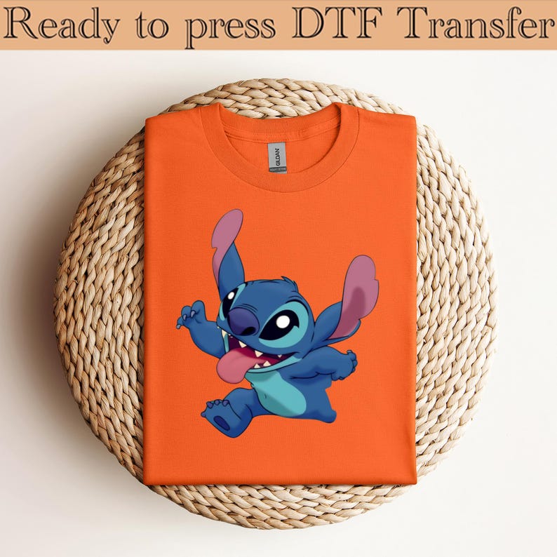 May include: Orange t-shirt featuring a cartoon character, Stitch, with blue fur, large ears, and a pink tongue. The text "Ready to press DTF Transfer" is at the top. The shirt is folded and placed on a woven, circular surface.