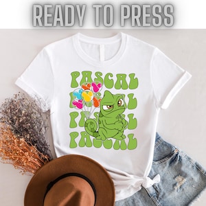 May include: White t-shirt with a green chameleon holding colorful balloons. The chameleon is looking to the right and has a grumpy expression. The text "PASCAL" is repeated in a retro font.