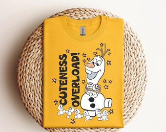 Puede incluir: Camiseta de color amarillo mostaza con un gráfico de Olaf de Frozen, rodeado de estrellas y las palabras "CUTENESS OVERLOAD!". La camiseta se muestra sobre un salvamanteles circular tejido. El texto "Ready to Press DTF Transfer" está en la parte superior.