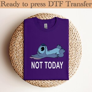 K&ouml;nnte beinhalten: Ein violettes Kurzarm-T-Shirt mit einer Grafik einer blauen au&szlig;erirdischen Figur, die auf dem R&uuml;cken liegt, darunter der Text NOT TODAY in wei&szlig;en Gro&szlig;buchstaben. Das T-Shirt hat einen Rundhalsausschnitt und ein sichtbares Markenetikett innen.