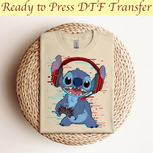May include: A beige t-shirt with a blue Stitch character wearing red headphones and holding a video game controller. The background has a glitch effect with blue and red lines.