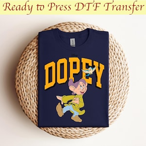 May include: A navy blue t-shirt with a yellow graphic of the Disney character Dopey from Snow White and the Seven Dwarfs. The graphic includes the word "Dopey" in yellow letters and a small blue bird flying above the character.