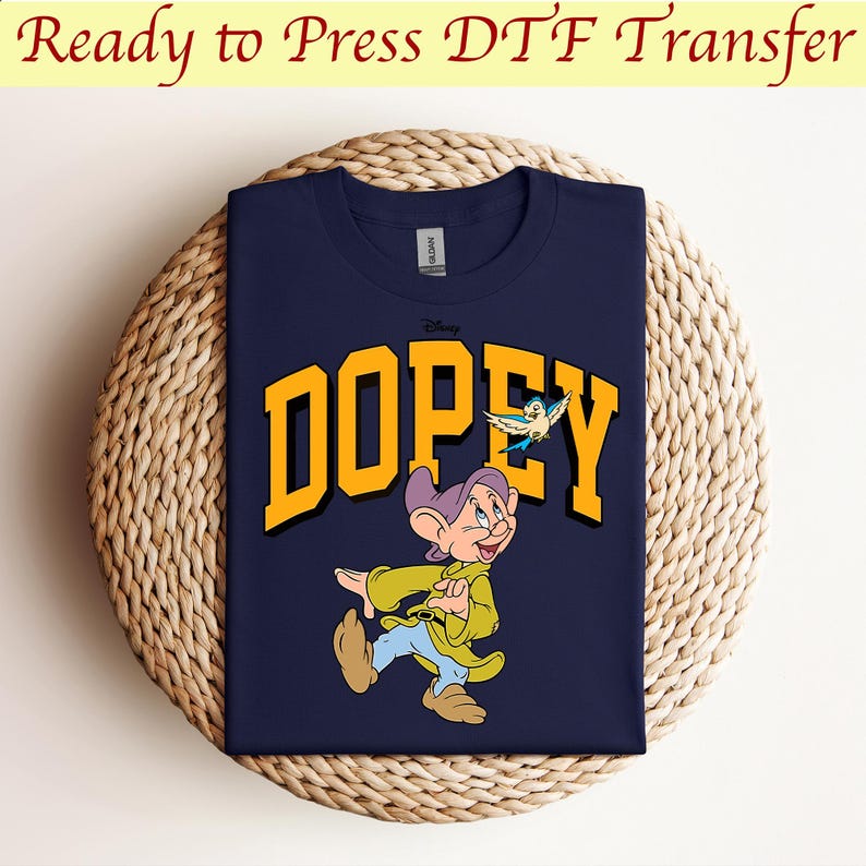 May include: A navy blue t-shirt with a yellow graphic of the Disney character Dopey from Snow White and the Seven Dwarfs. The graphic includes the word "Dopey" in yellow letters and a small blue bird flying above the character.