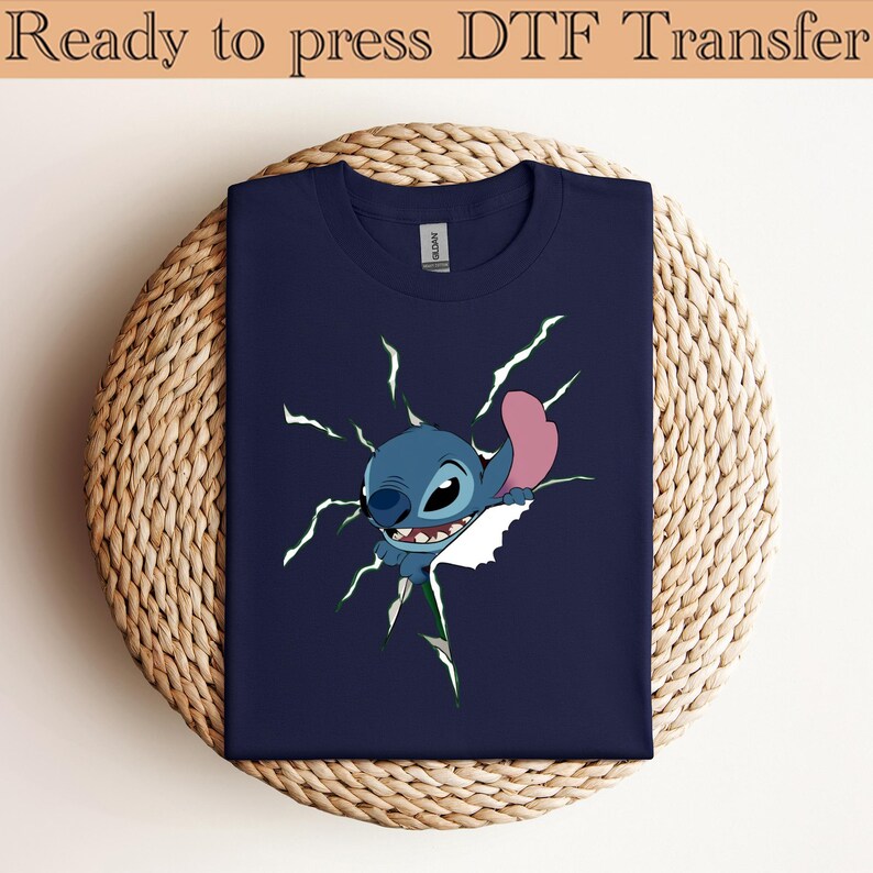 K&ouml;nnte beinhalten: Marineblaues T-Shirt mit einem Cartoon-Stitch-Design, das durch einen wei&szlig;en Riss mit gr&uuml;nen Blitzen bricht. Der Text "Ready to press DTF Transfer" steht oben. Das T-Shirt ist auf einer gewebten, kreisf&ouml;rmigen Oberfl&auml;che gefaltet.