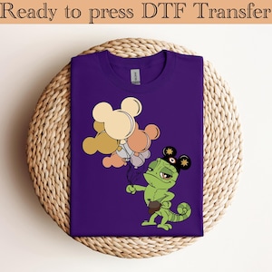 Könnte beinhalten: Lilafarbenes T-Shirt mit einem grünen Chamäleon, das einen Mickey-Mouse-Hut trägt und Mickey-Mouse-Ballons hält.