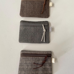 May include: Three rectangular zippered pouches in shades of brown and gray. The top pouch has a brown grid pattern, the middle is dark gray with small white dots, and the bottom features a brown checkered texture. Each has a side fabric tag.