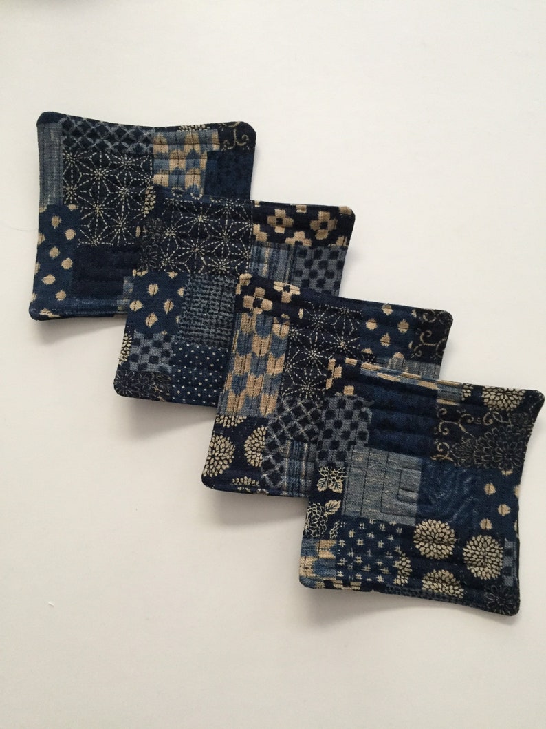 Może przedstawiać: Cztery niebieskie i kremowe podkładki pod kubki z pikowanego materiału w stylu patchwork. Podkładki są wykonane z bawełny i idealnie nadają się do ochrony mebli przed ciepłem i zarysowaniami.