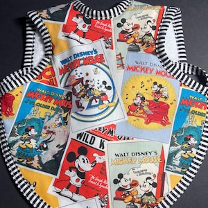 May include: A baby bib featuring a collage of vintage Walt Disney's Mickey Mouse comic book covers. The bib has a black and white striped trim and a white terry cloth backing. The bib is a colorful mix of red, yellow, blue, and white.