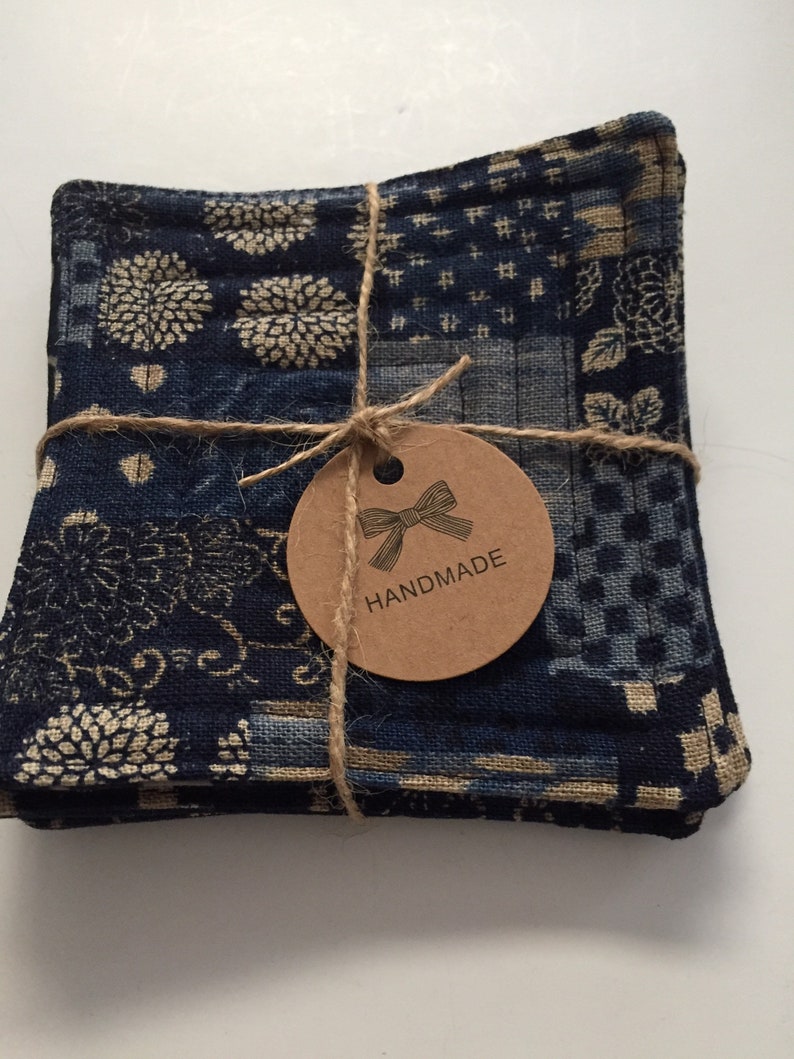 Może przedstawiać: Zestaw czterech niebieskich i białych podkładek pod kubki wykonanych z tkaniny w stylu patchwork. Podkładki są związane sznurkiem i ozdobione brązową etykietą z napisem "HANDMADE".