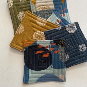 May include: A collection of five square fabric coasters featuring quilted stitching in concentric square patterns. The coasters display various colors including olive green, mustard yellow, and shades of blue, with printed floral and abstract shapes.