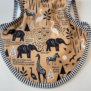 May include: A baby bib with a tan background featuring a black and white safari animal print. The print includes elephants, giraffes, zebras, birds, and trees. The bib has a black and white striped border.