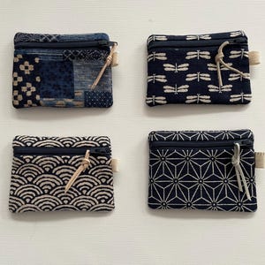 May include: Four rectangular fabric coin pouches with dark blue and cream patterns. Each pouch features a black zipper closure with a light-colored leather pull tab and a small side loop. The designs include patchwork, dragonflies, waves, and geometric stars.
