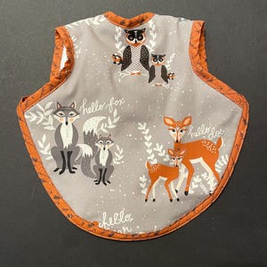 May include: A gray baby bib with an orange trim and a woodland animal print. The bib features illustrations of foxes, owls, and deer, with the words "hello fox" and "hello deer" printed on the front.