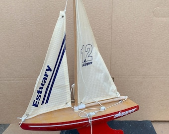 Puede incluir: Un velero de juguete de madera rojo y blanco con una vela blanca que dice "12 skipper" y "Estuary". El velero está en un soporte de madera.