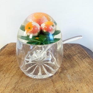 Può includere: Una zuccheriera di vetro trasparente con un coperchio a cupola che presenta un design realistico di frutta di arancia. Il coperchio ha tre arance e foglie verdi. La ciotola ha un cucchiaio di vetro trasparente.