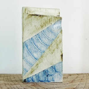 Könnte beinhalten: Eine kleine, rechteckige Keramikvase mit einem geometrischen Muster in Blau und Weiß. Die Vase hat eine raue, strukturierte Oberfläche und einen leicht unebenen oberen Rand.