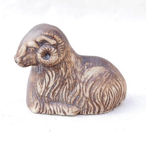 Puede incluir: Figura de carnero de cerámica en posición sentada. La escultura es principalmente marrón con acentos beige más claros, mostrando texturas detalladas de pelaje y cuernos rizados. La cabeza del carnero está ligeramente girada hacia la izquierda.