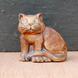 Puede incluir: Figura de gato de cerámica marrón. El gato está sentado con la cola enrollada alrededor de su cuerpo. La figura tiene ojos grandes y redondos y bigotes detallados. Está sobre una superficie de madera, con un fondo gris oscuro.