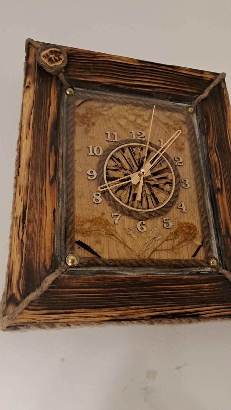 May include: A rustic wooden clock with a rope border and a natural wood face. The clock hands are gold and the numbers are black. The clock is set against a white wall.