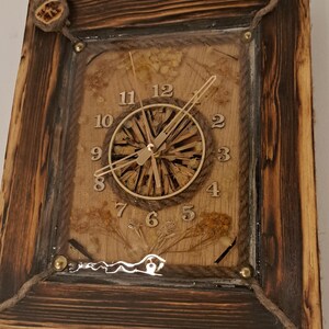 May include: A rustic wooden clock with a natural wood face and gold-coloured hands. The clock face is surrounded by dried flowers and plants, encased in clear resin. The clock is framed in a dark brown wooden frame with rope accents.