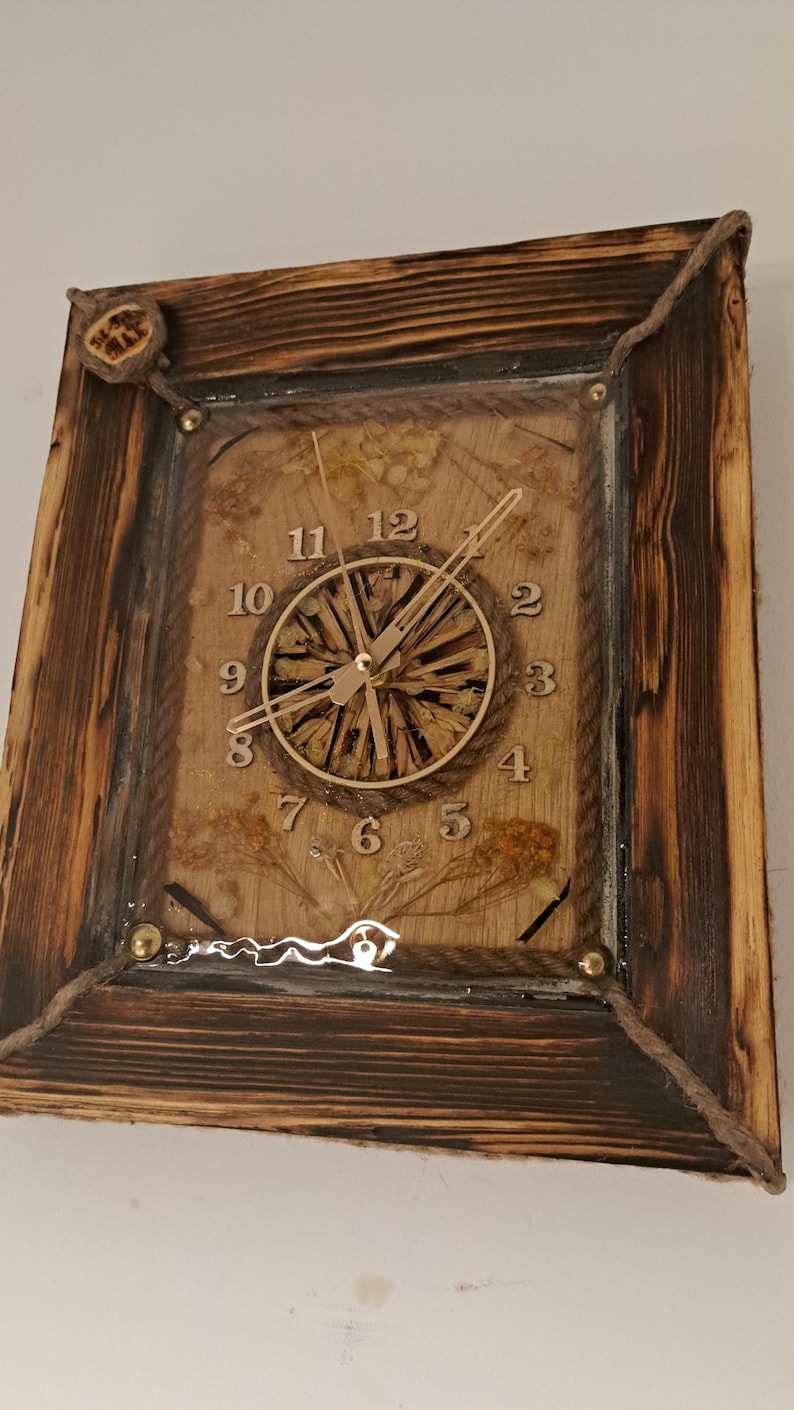 May include: A rustic wooden clock with a natural wood face and gold-coloured hands. The clock face is surrounded by dried flowers and plants, encased in clear resin. The clock is framed in a dark brown wooden frame with rope accents.
