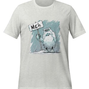 May include: A gray t-shirt with a cartoon yeti holding a sign that says "MEH". The yeti is white and furry with a grumpy expression. The background is a blue and white snowy scene.