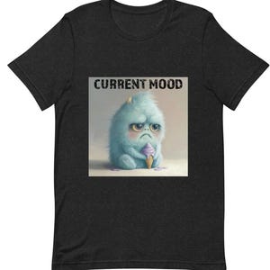 Op de afbeelding: Een donkergrijs T-shirt met een blauw en wit cartoonmonster dat een ijsje vasthoudt. De tekst "CURRENT MOOD" is boven het monster gedrukt.