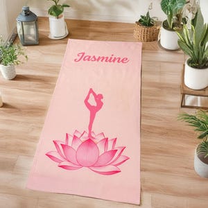 May include: Pink yoga mat featuring a lotus flower design and a yoga pose silhouette. The name "Jasmine" is printed at the top. The mat is displayed on a wooden floor.